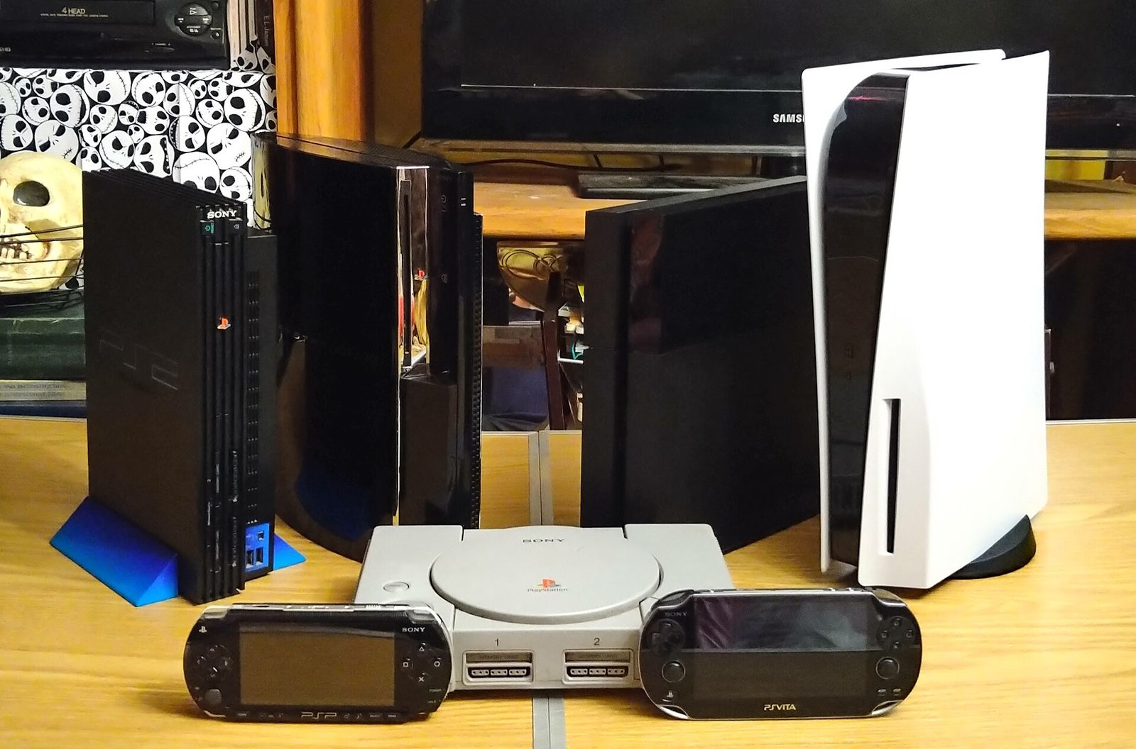 桌上並排的歷代 PlayStation 主機，從灰色的初代、黑色的第 2 到第 4 代，到白色直立的 PS5，前面放著兩台掌機