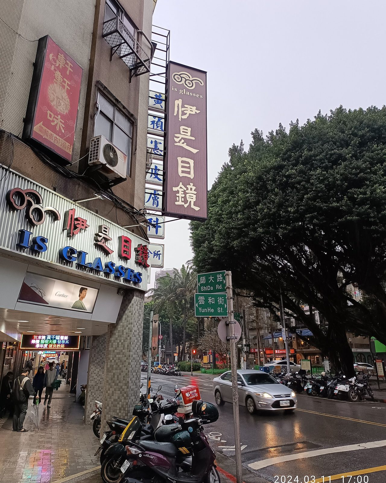 下雨天傍晚的台北師大路口，轉角騎樓上掛著直式與橫式的眼鏡行招牌，路口停著機車與汽車