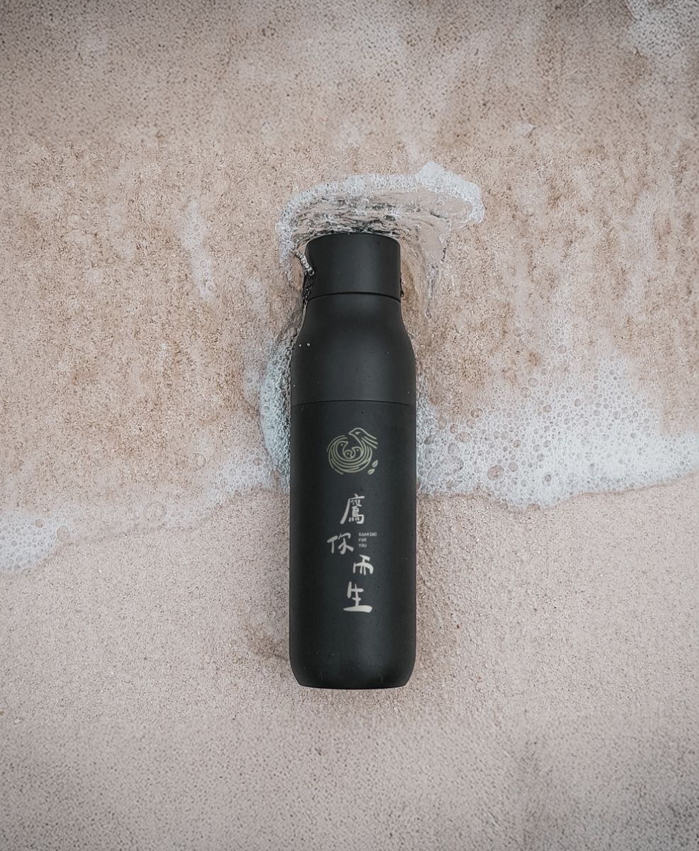浪花沙灘上的黑色隨行瓶，瓶身直印鷹你而生 Logo 與字