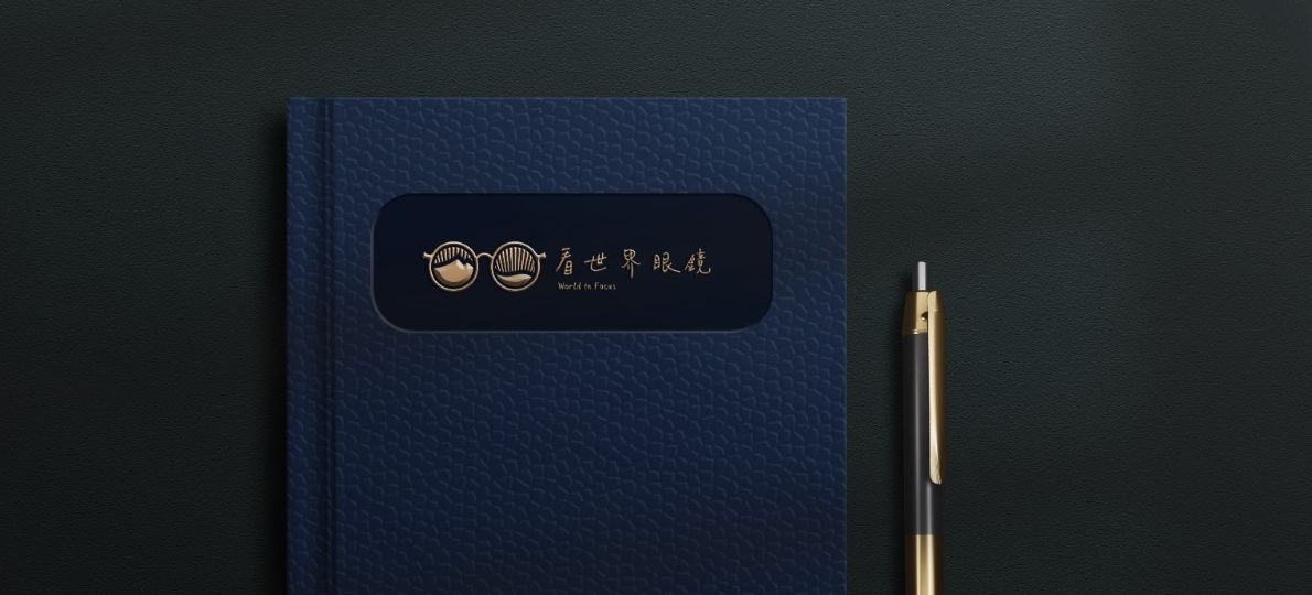 深藍皮革筆記本封面嵌著金色看世界眼鏡標誌，旁邊一支金色原子筆