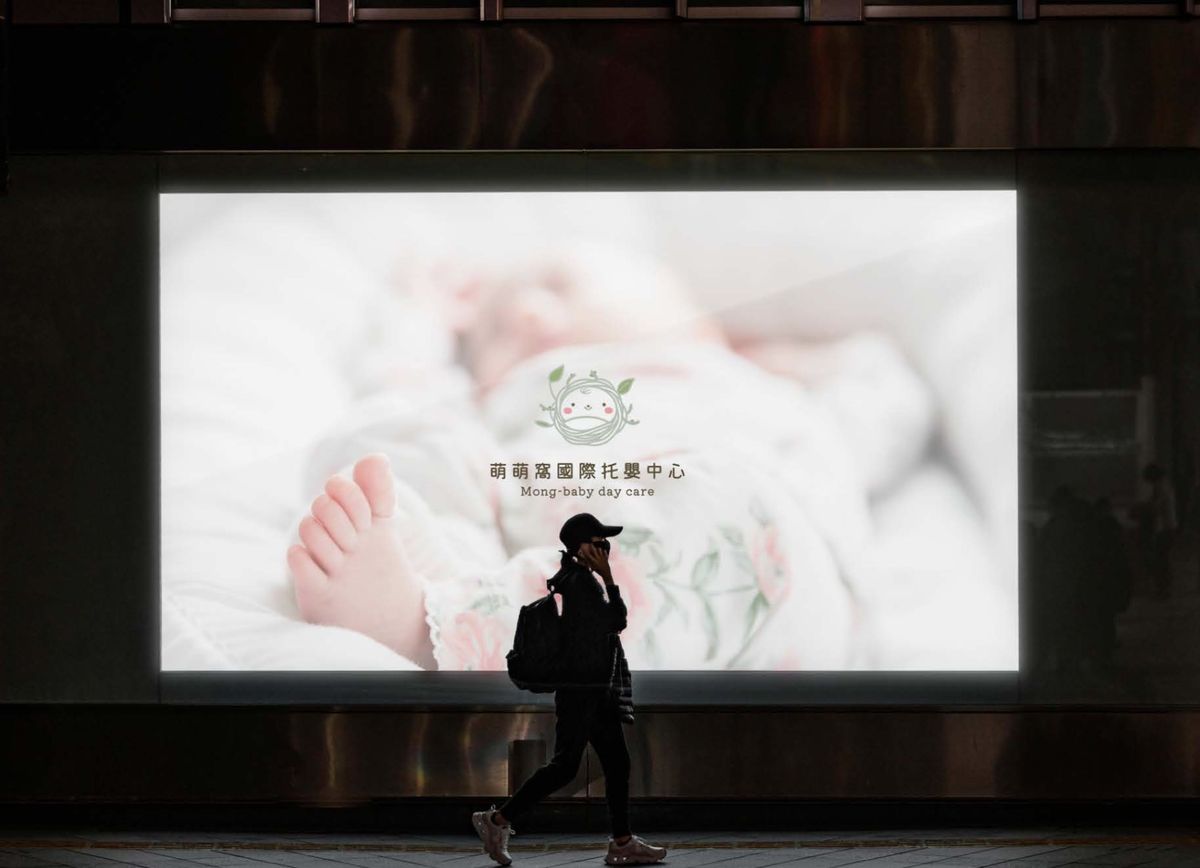夜晚街邊的大型發光看板，畫面是包巾裡的嬰兒小腳與萌萌窩標誌，前方有行人走過