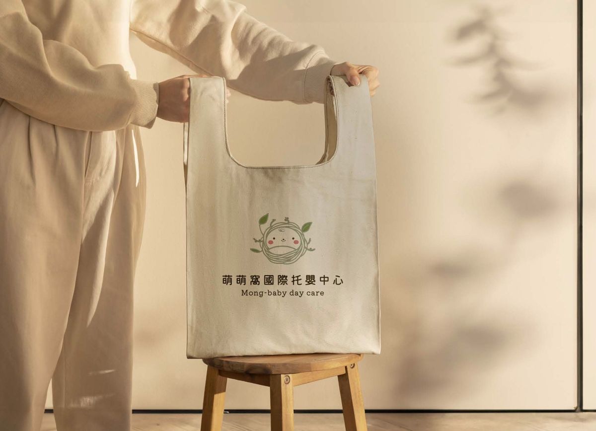 雙手提起放在木凳上的米白帆布提袋，印著萌萌窩標誌