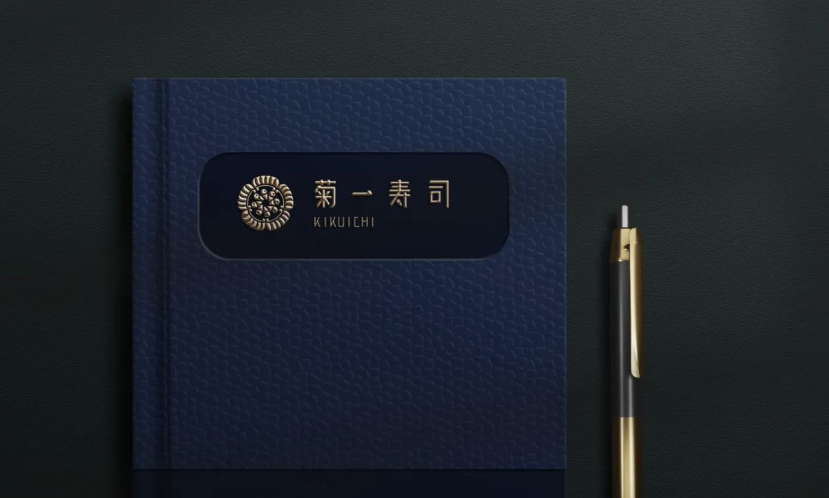 深藍色筆記本封面嵌著金色的菊一寿司標誌，旁邊放著一支金色的筆